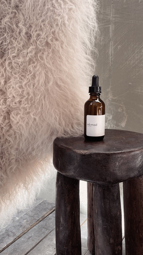 Nomad Room Oil amber glass bottle styled on dark wood stool in warm interior setting.