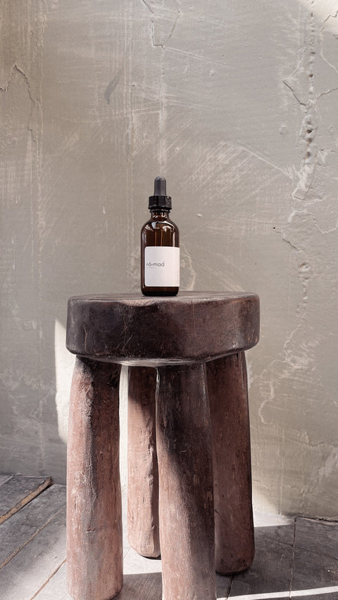 Nomad Room Oil amber glass bottle styled on dark wood stool in warm interior setting.