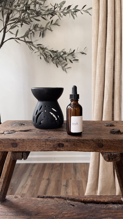 Souk Room Oil amber glass bottle styled with black ceramic oil burner on rustic wood table.