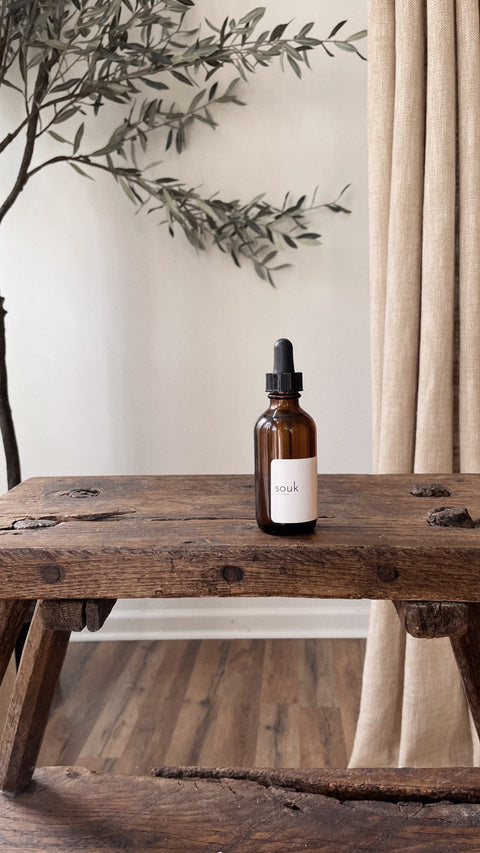Souk Room Oil amber glass bottle styled on rustic wood table with branches in warm interior setting.