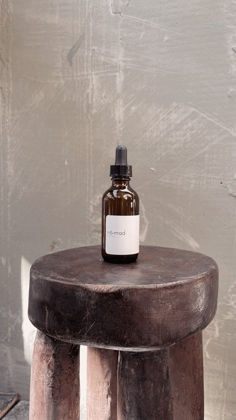 Nomad Room Oil amber glass bottle styled on dark wood stool in warm interior setting.
