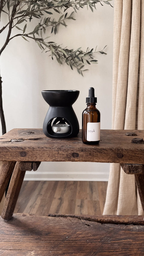 Souk Room Oil amber glass bottle styled with black ceramic oil burner on rustic wood table.