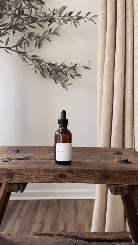 Souk Room Oil amber glass bottle styled on rustic wood table with branches in warm interior setting.